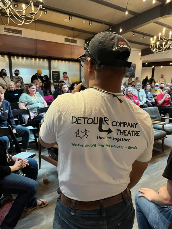 Seen from behind, a storyteller in a cap and a Detour Company Theatre T-shirt speaks into a microphone to a full room of seated audience members.