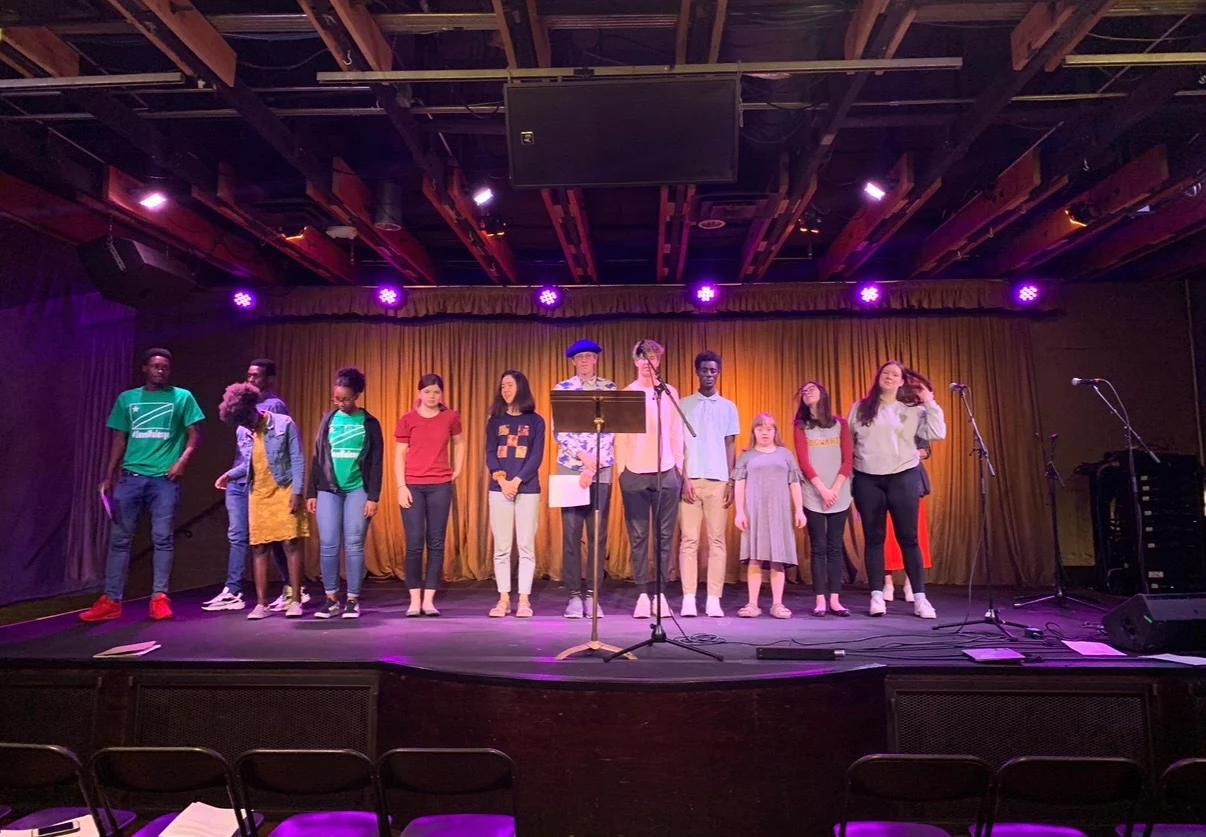 A group of storytellers stands together in a line across a stage lit in purple and gold, a music stand and microphones in front of them.