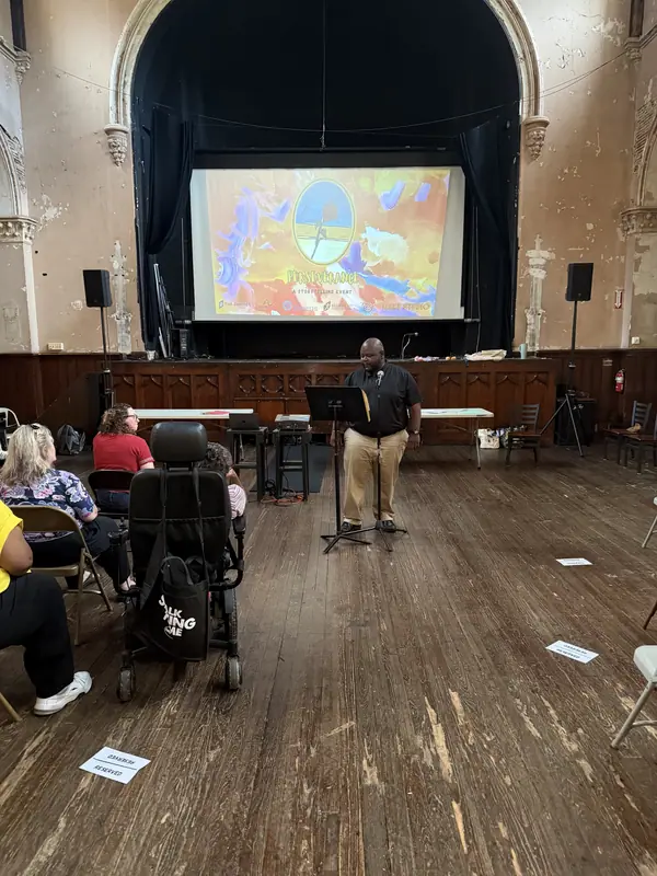 A storyteller rehearses at a music stand on stage, the event artwork projected behind him.